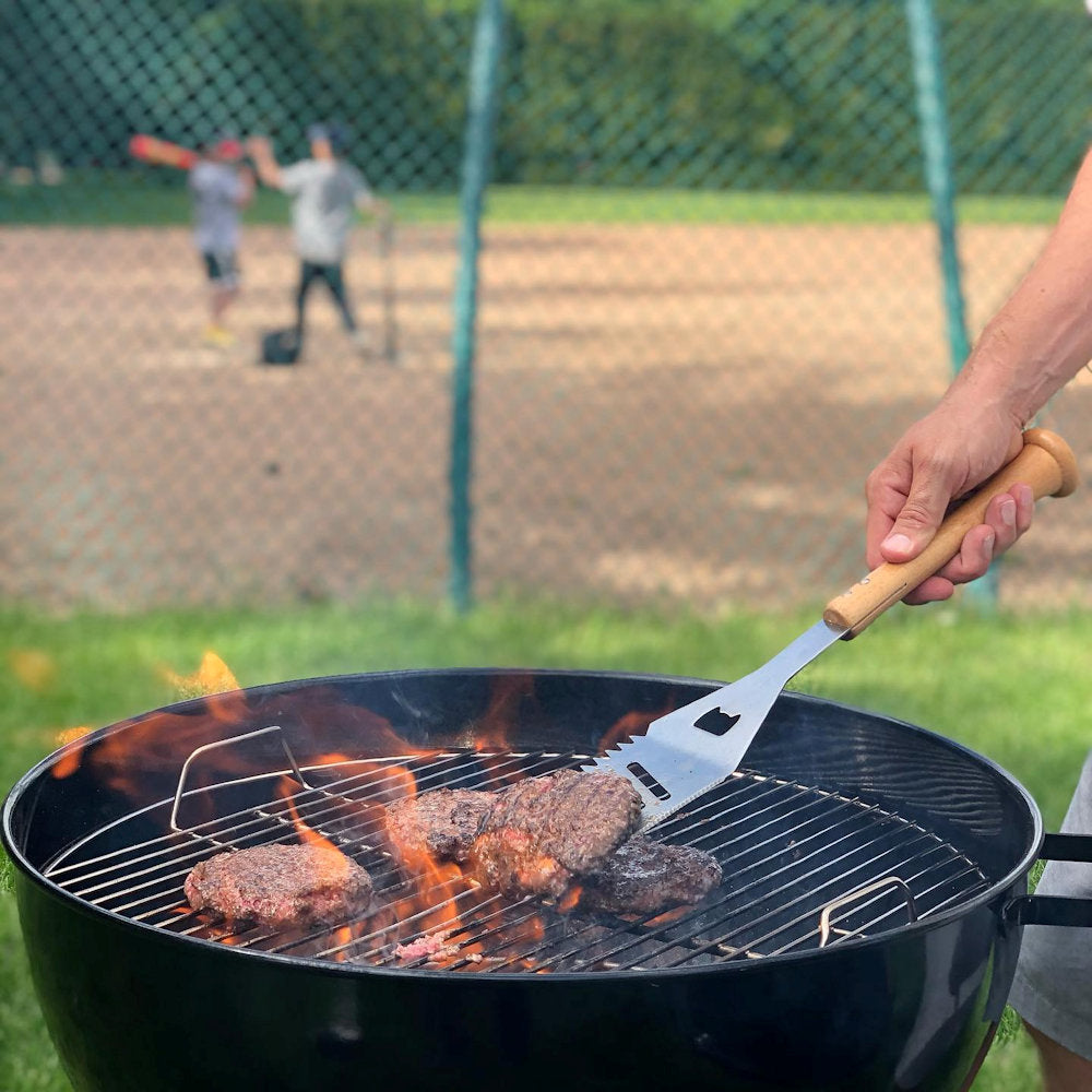 Person grilling hamburgers with a baseball spatula on a small barbecue with a baseball game in the background
