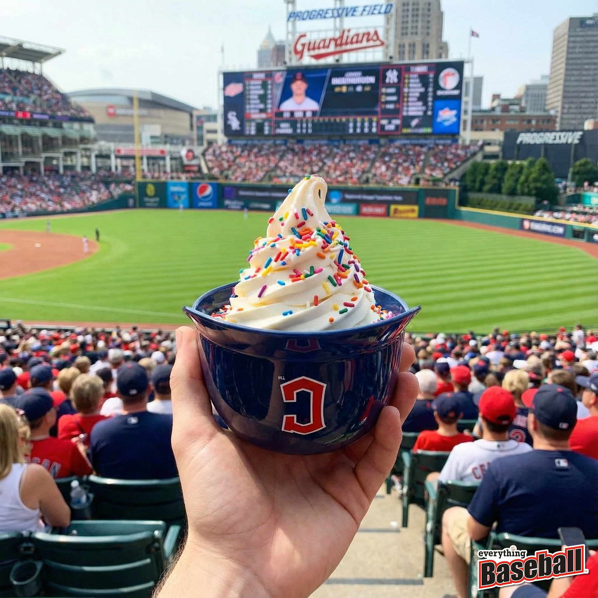 Ice cream helmet cup with colorful sprinkles held up at a Cleveland Guardians baseball game, with a large scoreboard and stadium in the background.