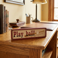 Wooden mini baseball sign with 'Play ball!' text on a desk with books and a lamp in the background