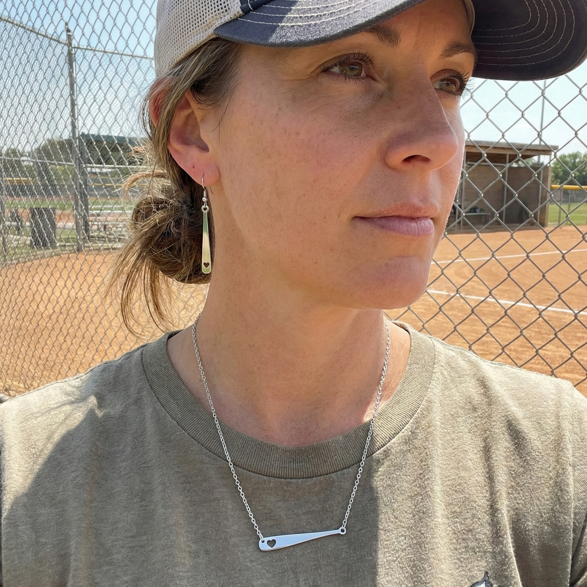 Woman showing off silver baseball bat heart necklace and earrings on a sports field wearing a cap and gray shirt