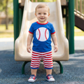 Child wearing a blue shirt with a baseball design and red and white striped shorts on a playground.