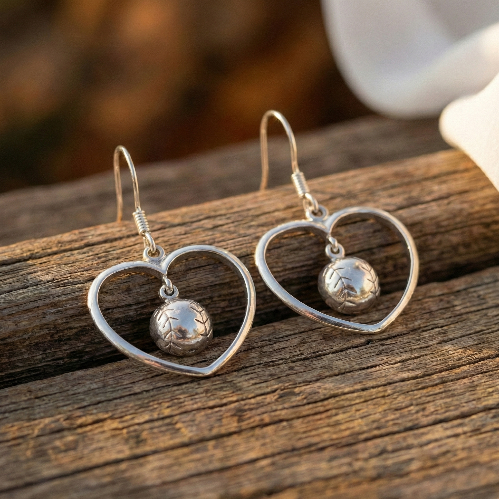 Silver heart-shaped earrings with a small baseball pendant on a wooden surface.