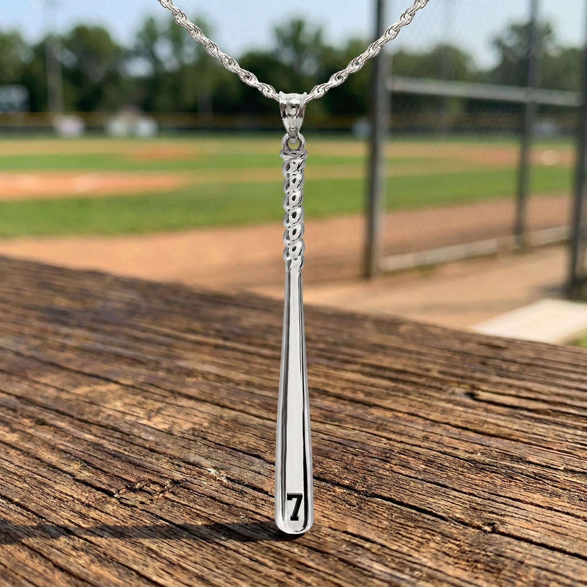 Silver necklace with a custom baseball bat number pendant on a wooden surface with a baseball field background