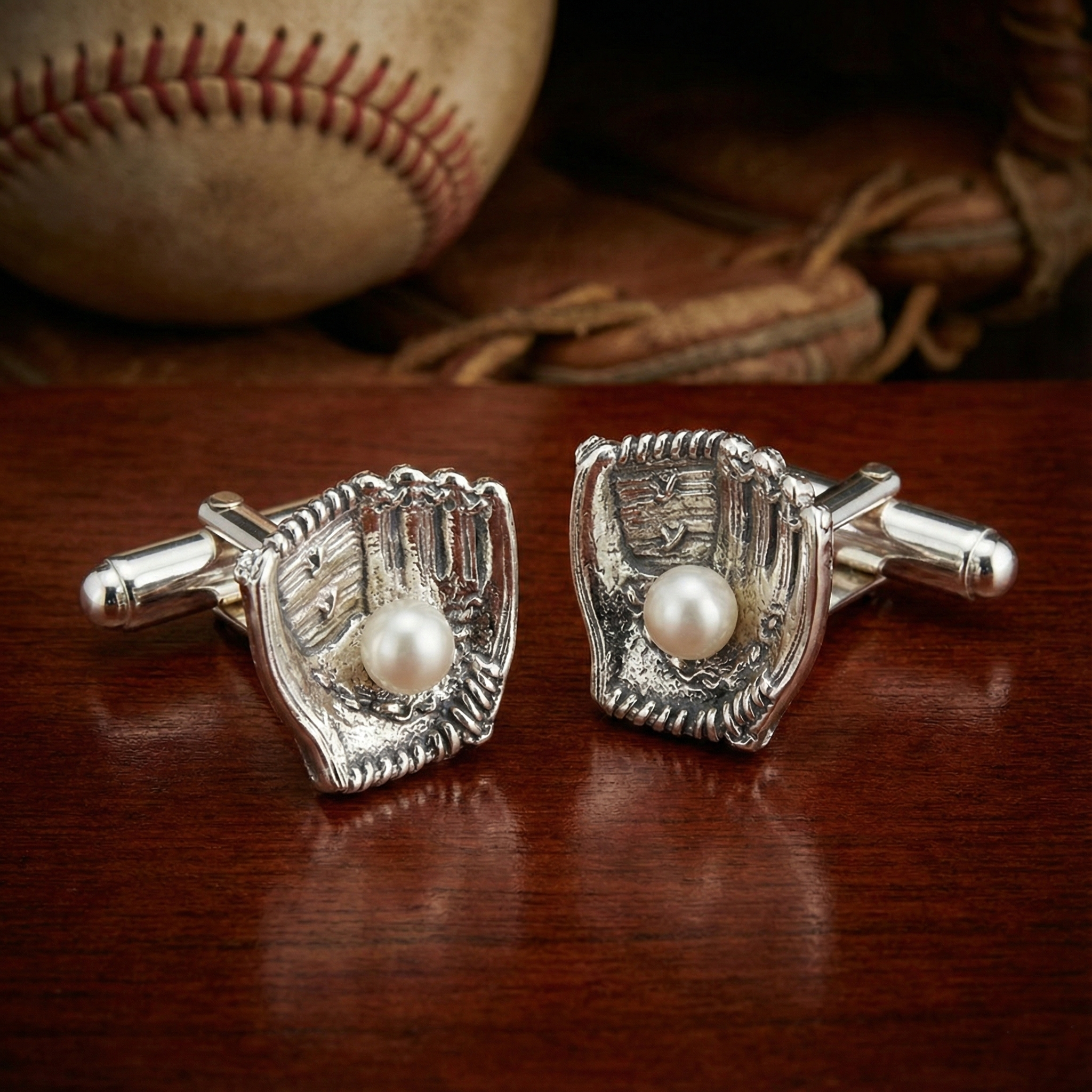 Silver baseball glove cufflinks with pearls on a wooden surface with a baseball and glove in the background.