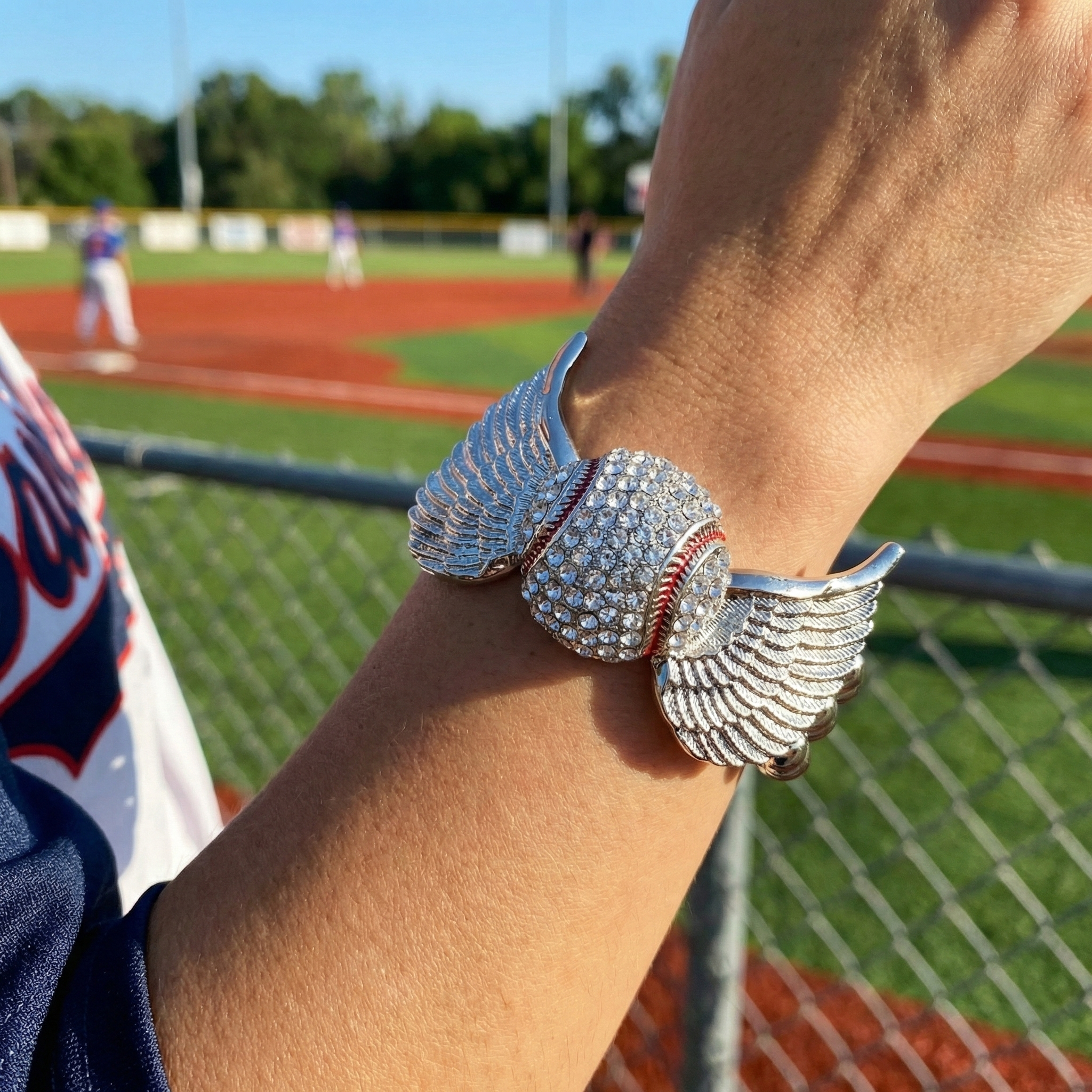 Close-up of a person's arm wearing a silver baseball wings stretch bracelet with a baseball field in the background