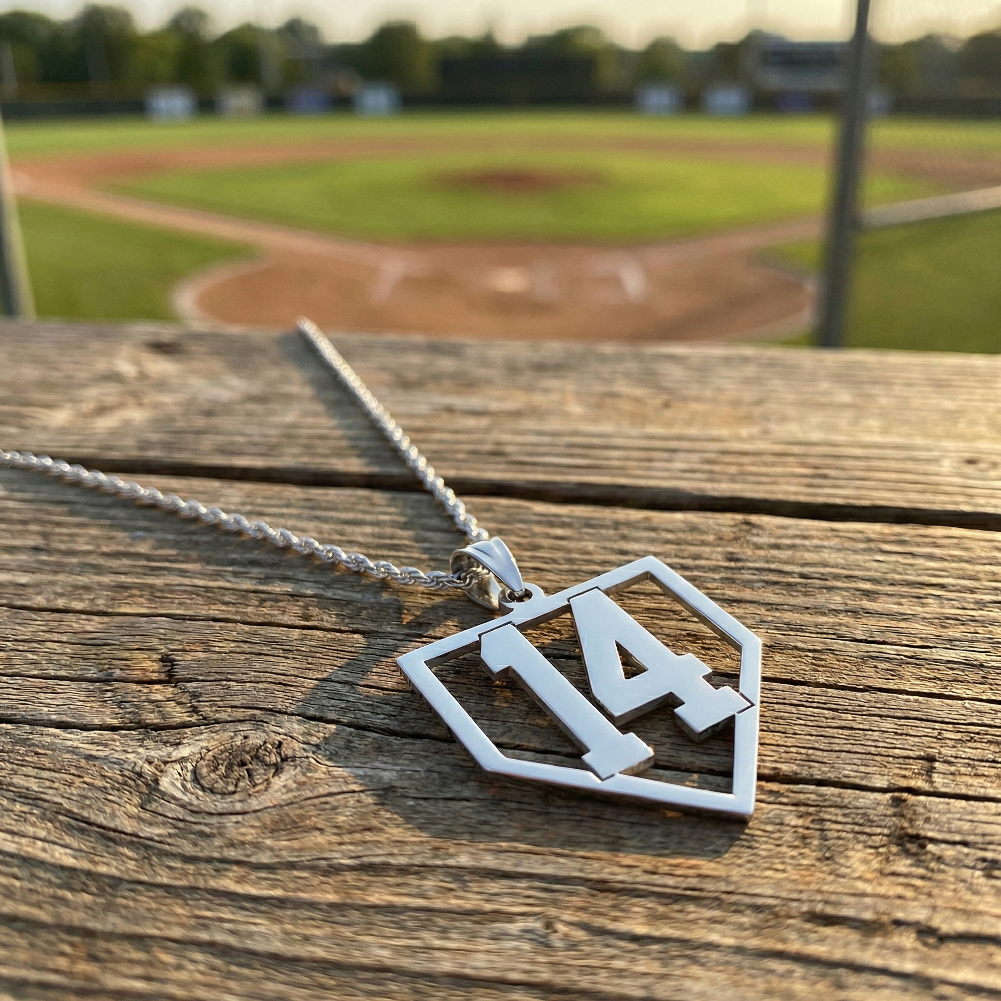 Silver baseball number home plate necklace with a pendant on a wooden surface, baseball field in the background