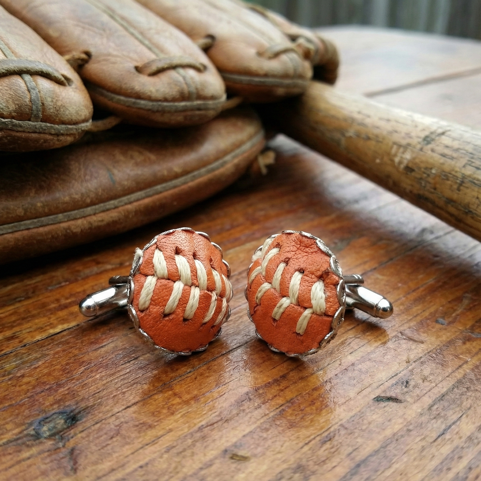 Pair of orange leather baseball cufflinks resembling a football on a wooden surface with a baseball glove and bat in the background.