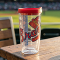 Clear tumbler with red lid featuring Atlanta Braves logo on a wooden table with a blurred baseball field background
