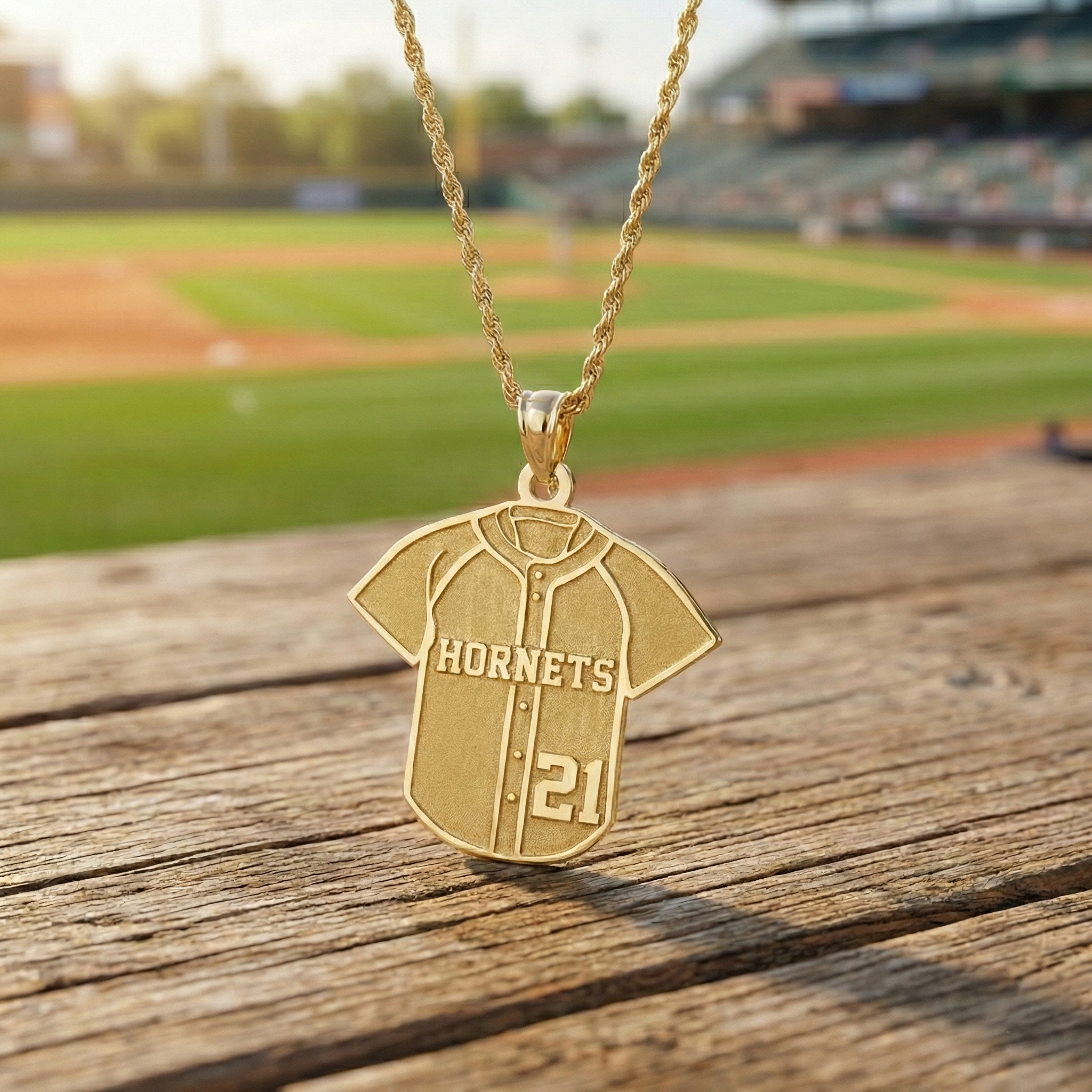 Gold 'Hornets' baseball jersey necklace with number 21 on a wooden surface with a baseball field background