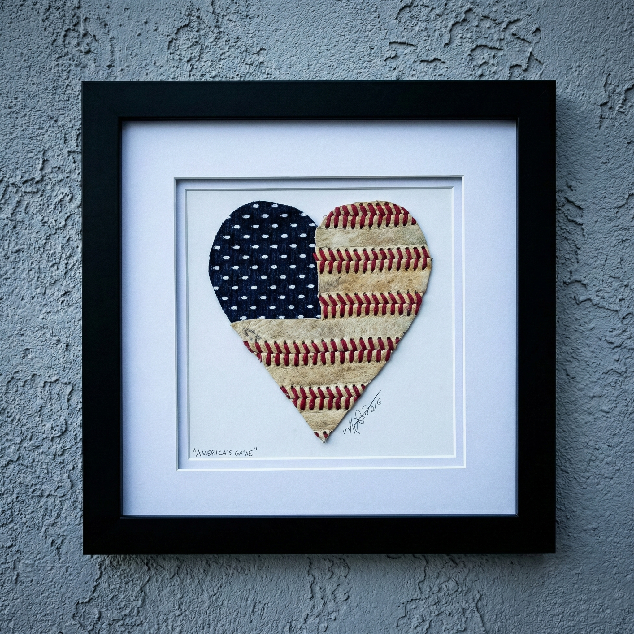 Framed artwork of a heart-shaped American flag made from baseballs on a textured gray wall.