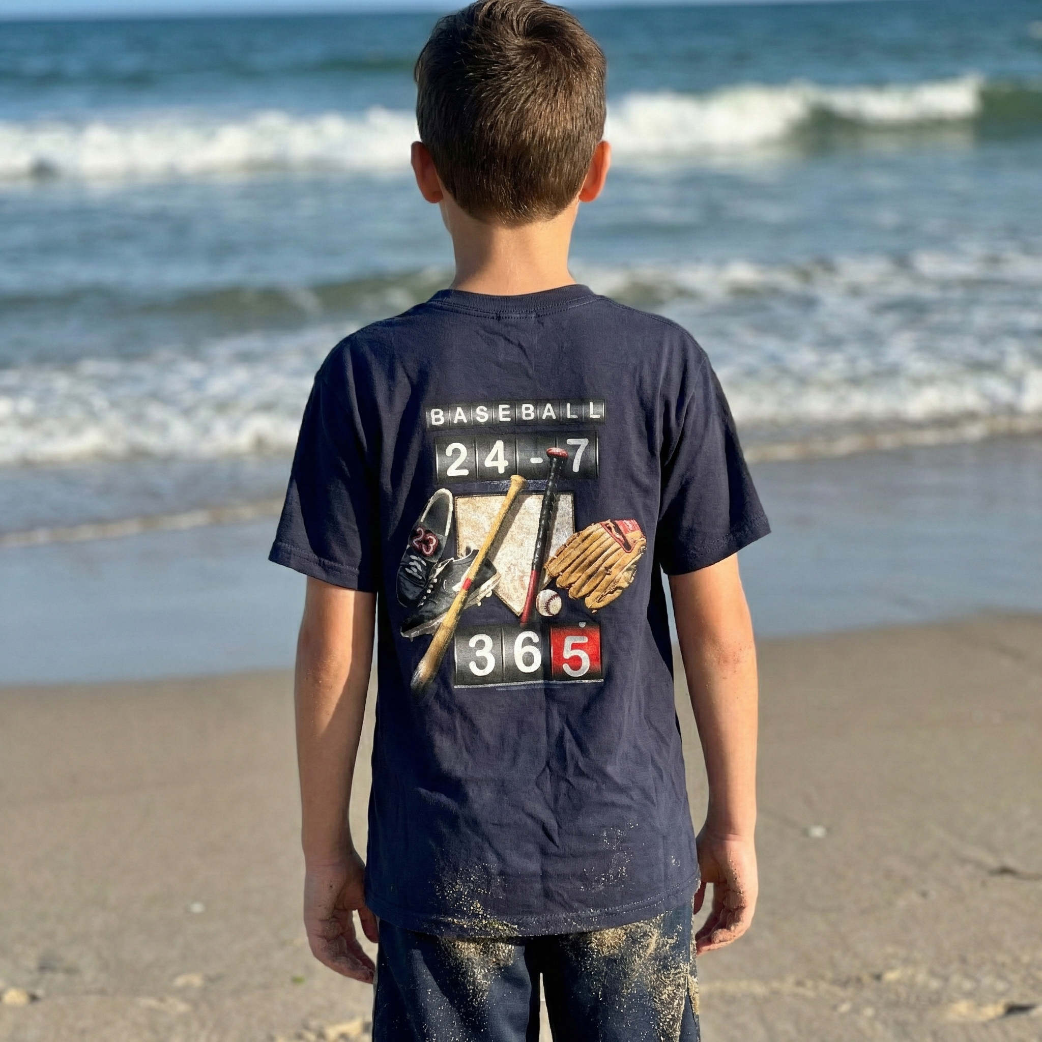 Child wearing a navy blue 24-7-365 baseball t-shirt with sports-themed design on a beach