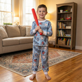 Child in baseball pajamas holding a red bat in a living room.