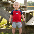 Child wearing a red and navy baseball shirt with gray shorts standing outdoors near wooden steps.