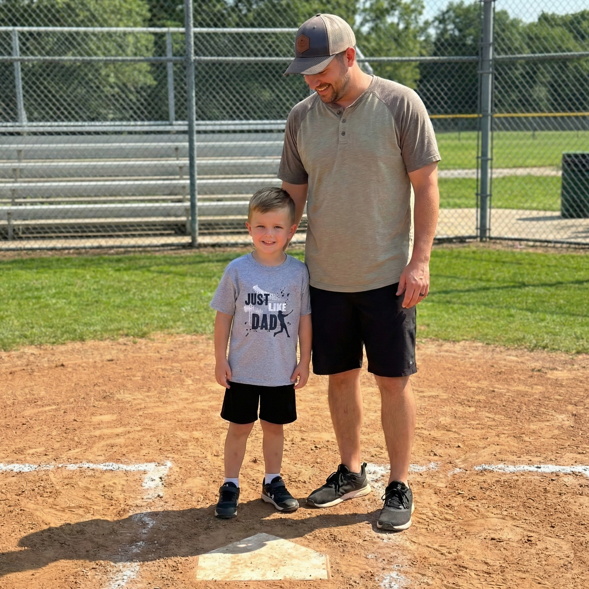 Man and child wearing Just Like Dad baseball t-shirt standing on a baseball field with a fence and grass in the background