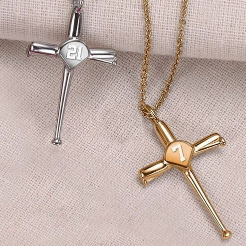 Silver and gold baseball cross necklaces with engraved numbers on a textured beige surface