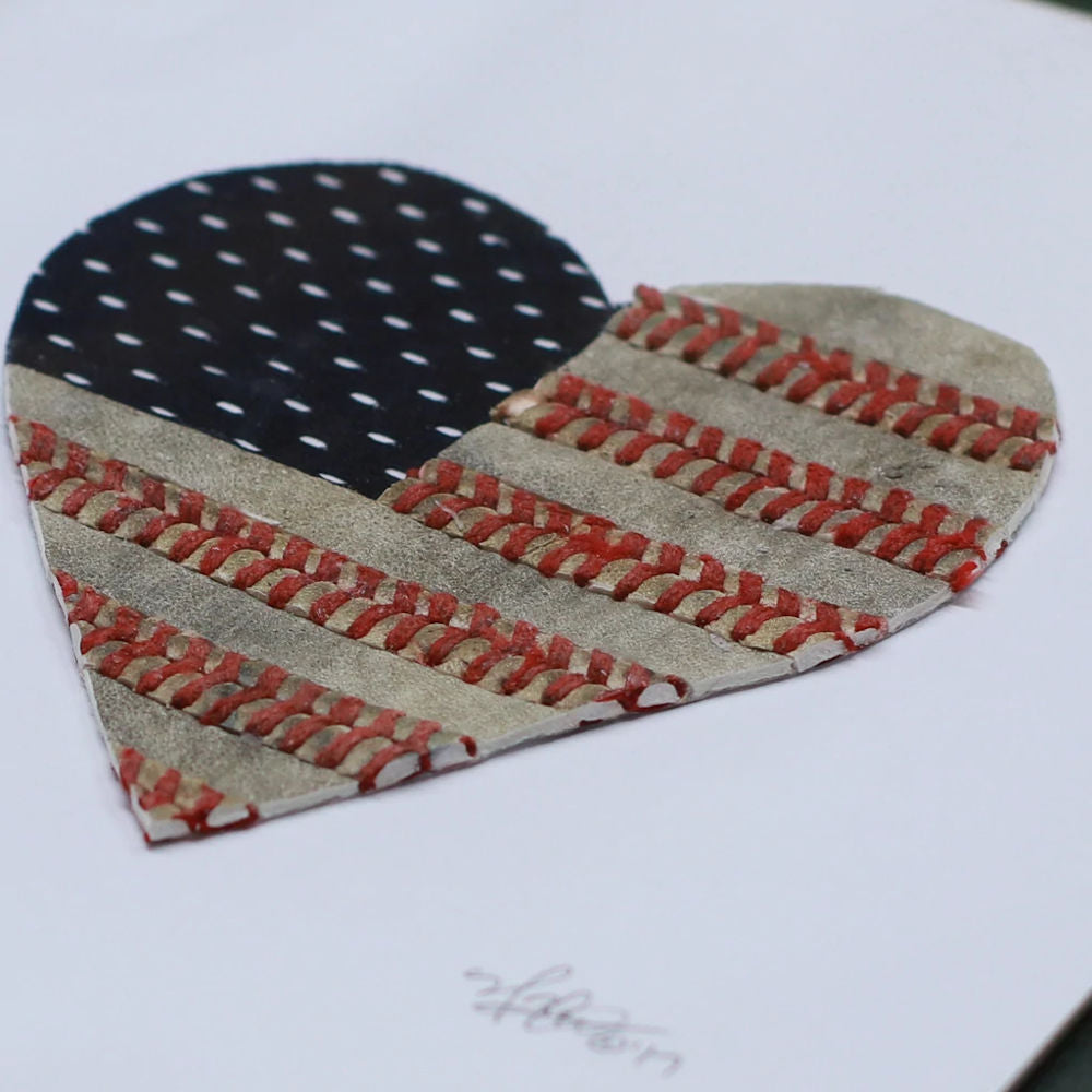 Heart-shaped American flag art made with blue jersey and read used baseball seams on a white board.