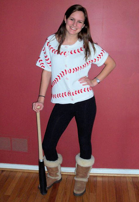 Person wearing a baseball-themed sweater, black pants, and brown boots holding a baseball bat against a red wall.