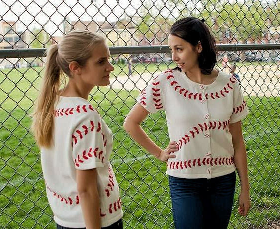 Two women wearing white sweaters with red baseball-stitch patterns standing in front of a chain-link fence.