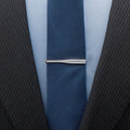Close-up of a navy tie with a silver baseball bat tie clip on a light blue shirt, worn with a black pinstripe suit.