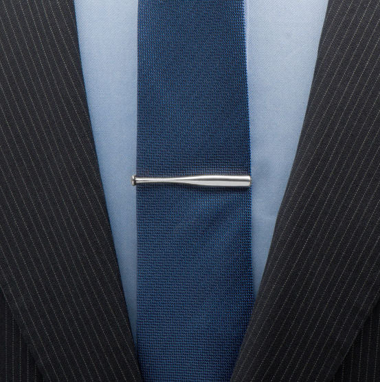 Close-up of a navy tie with a silver baseball bat tie clip on a light blue shirt, worn with a black pinstripe suit.