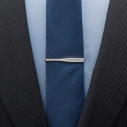 Close-up of a navy tie with a silver baseball bat tie clip on a light blue shirt, worn with a black pinstripe suit.