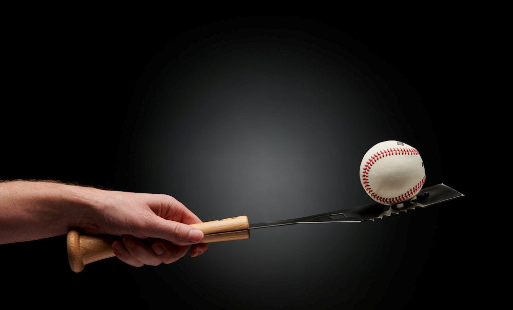 Hand holding a baseball bat grill spatula balancing a baseball on a black background