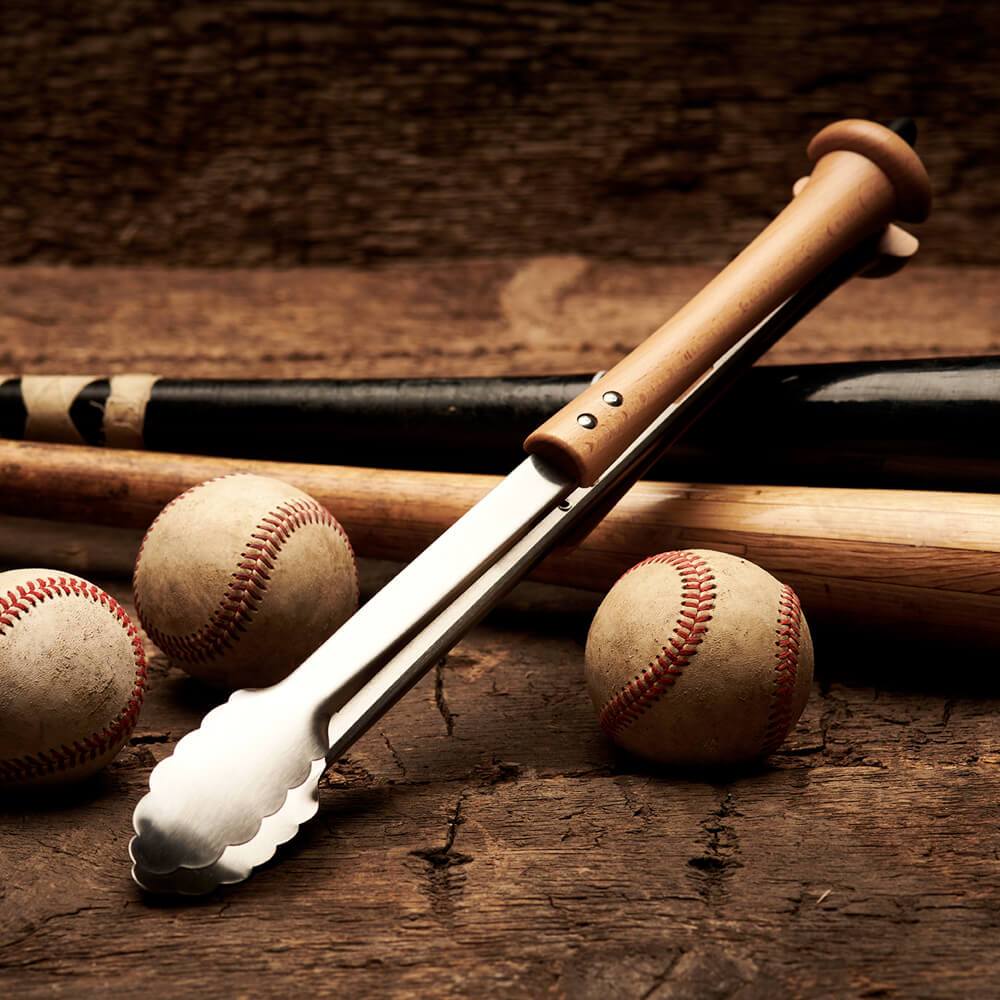 Metal grill tongs with wooden baseball bat handle on a wooden surface with baseballs and bats.