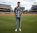 Man standing on a baseball field wearing a patterned shirt and jeans.