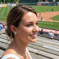 Woman with crystal baseball heart post earrings smiling at a baseball game with a field and spectators in the background