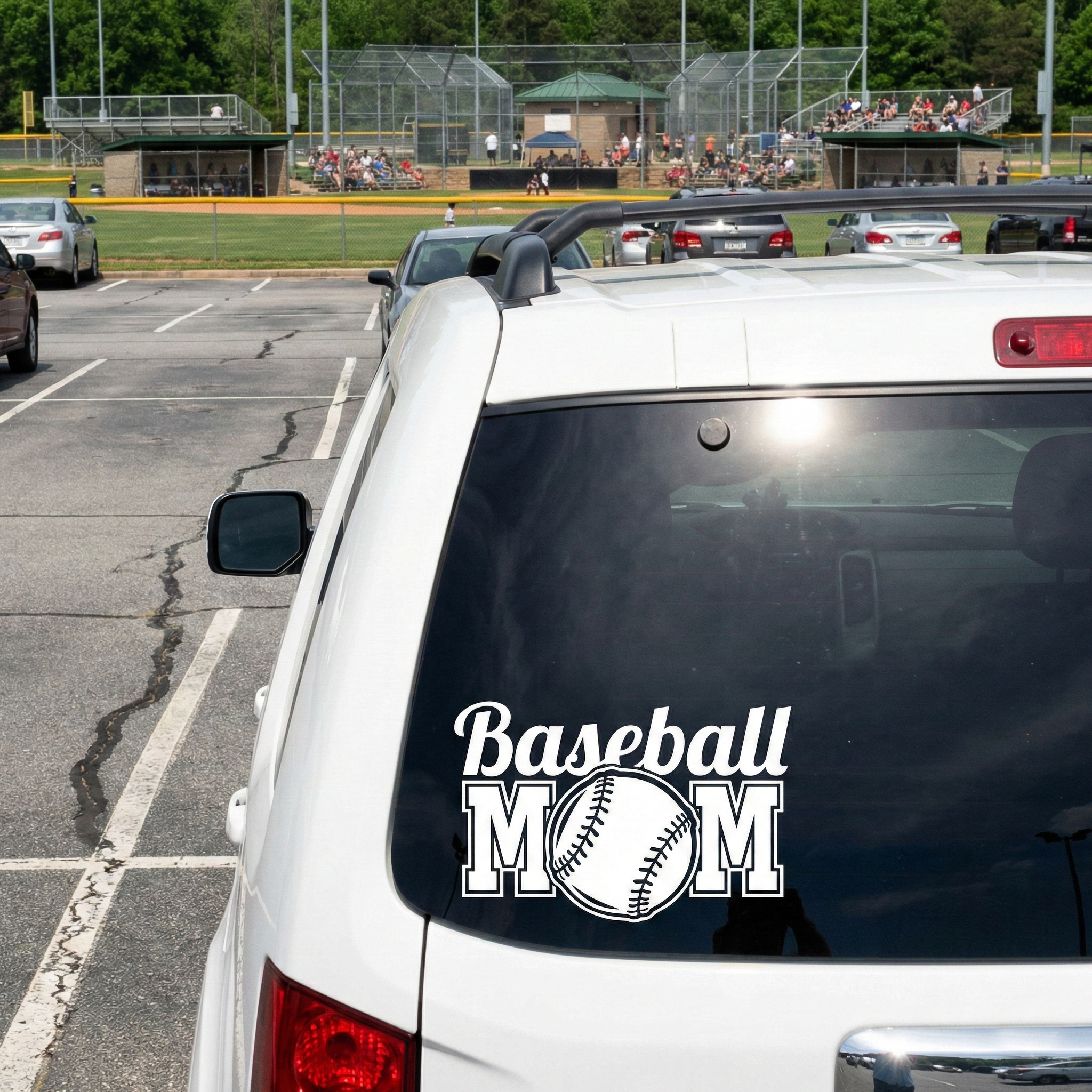 White car with 'Baseball Mom' decal in a parking lot near a baseball field.
