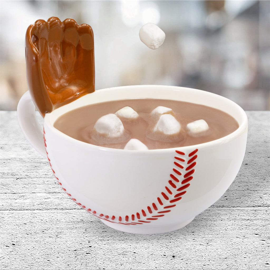 White baseball mug with a glove bowl with red design filled with hot chocolate and marshmallows, featuring a brown hand-shaped object on a light wooden surface.