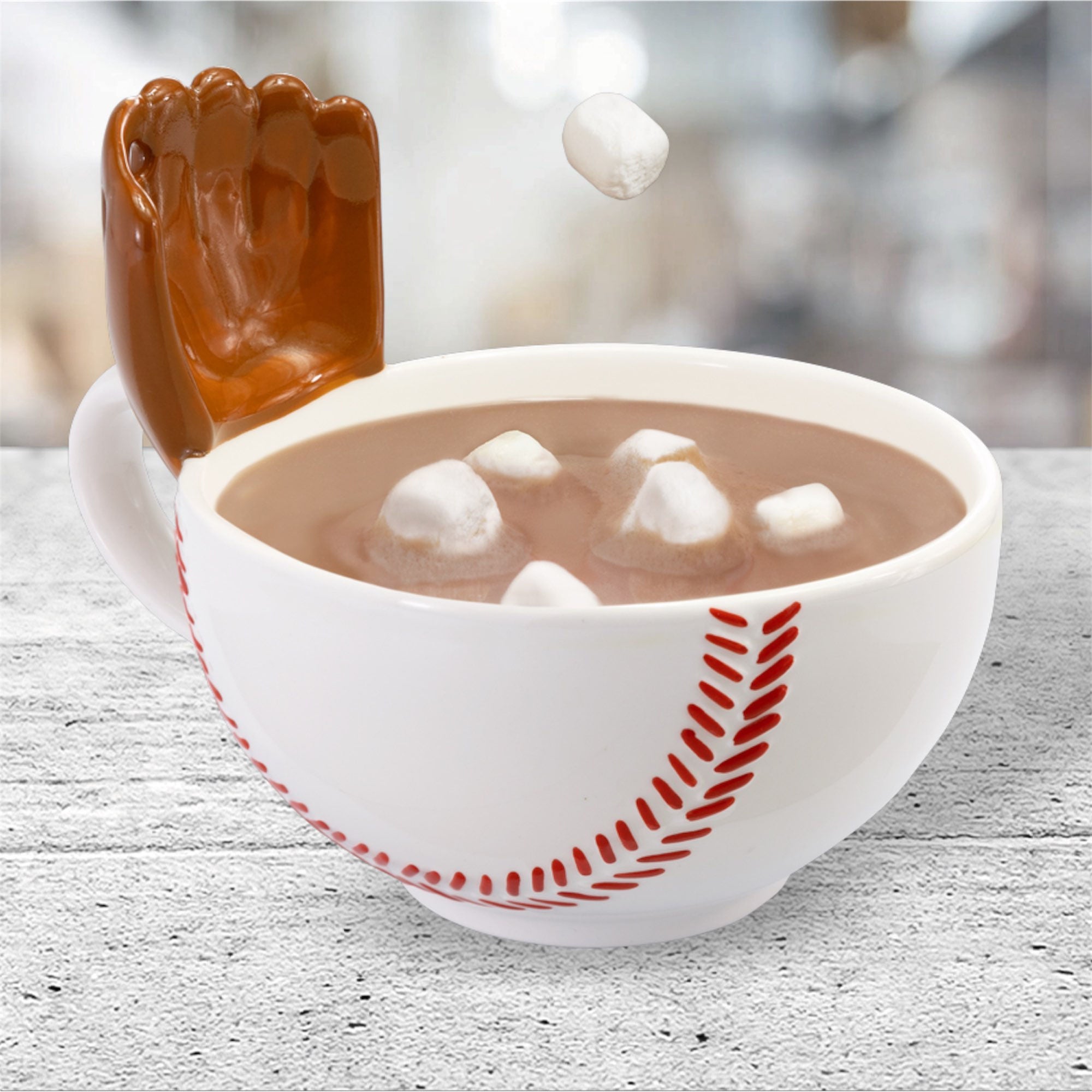 White baseball mug with a glove bowl with red design filled with hot chocolate and marshmallows, featuring a brown hand-shaped object on a light wooden surface.