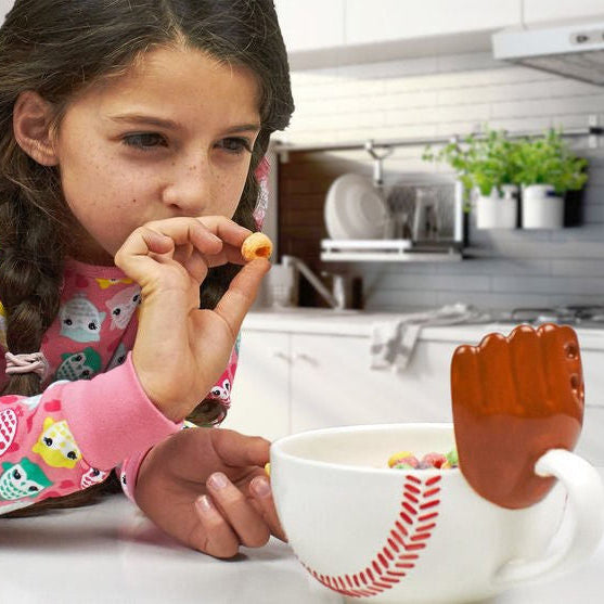 Young girl shooting cereal into her baseball mug with a glove in a kitchen setting