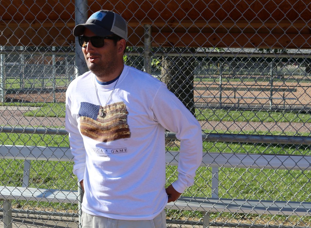 Man wearing a white long-sleeve shirt with a design, standing in front of a chain-link fence.