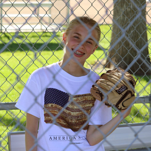Person wearing a white t-shirt with an American flag design, holding a baseball glove behind a chain-link fence.