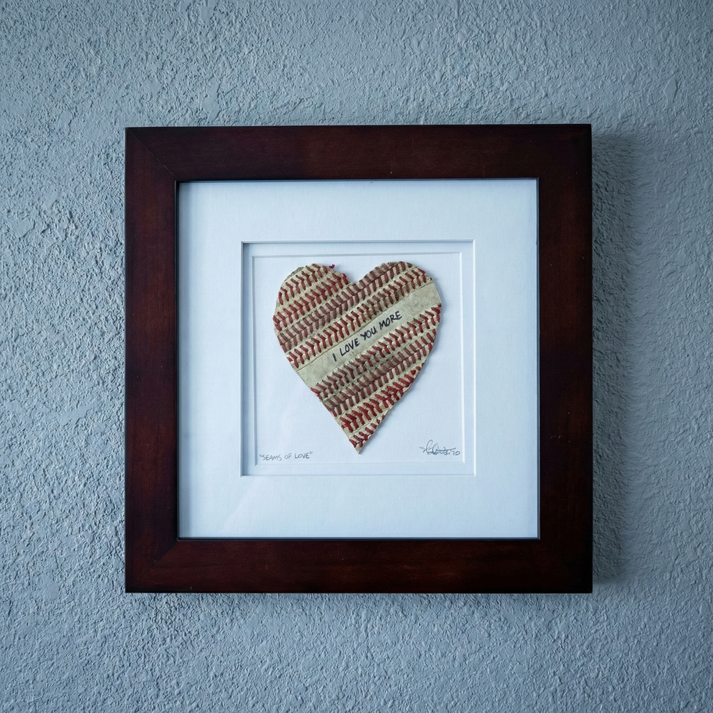 Framed heart-shaped baseball seams artwork with 'I Love You More' text on a blue wall