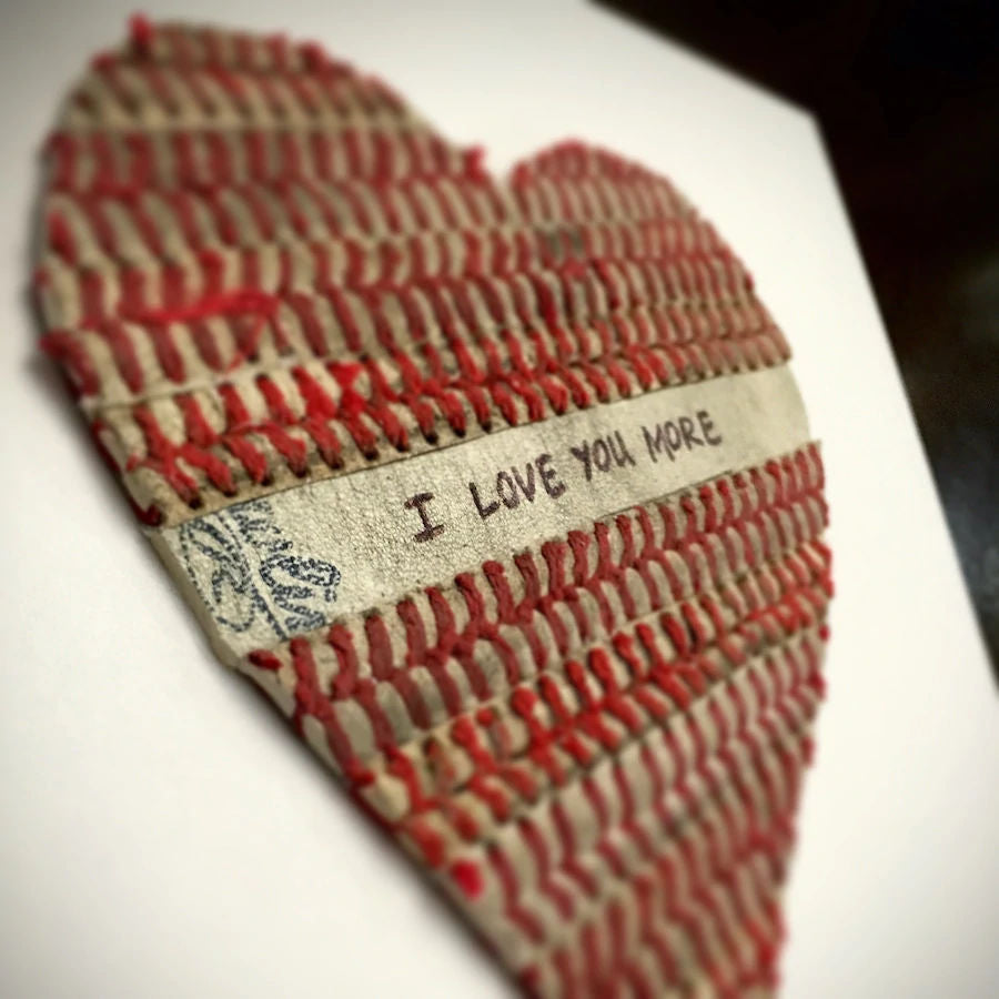 Heart-shaped baseball seams art with 'I Love You More' text on a white background