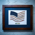 Framed artwork of an American flag made from baseballs on a textured blue wall.
