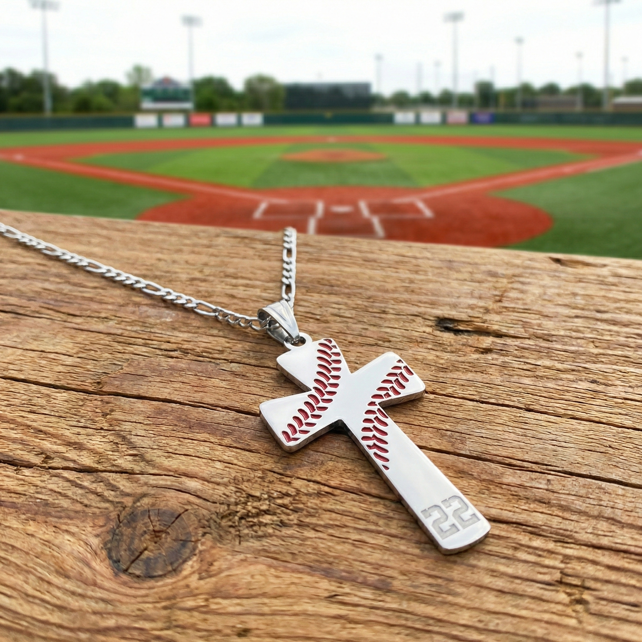 Silver baseball number cross pendant with baseball design on a wooden surface with a baseball field in the background