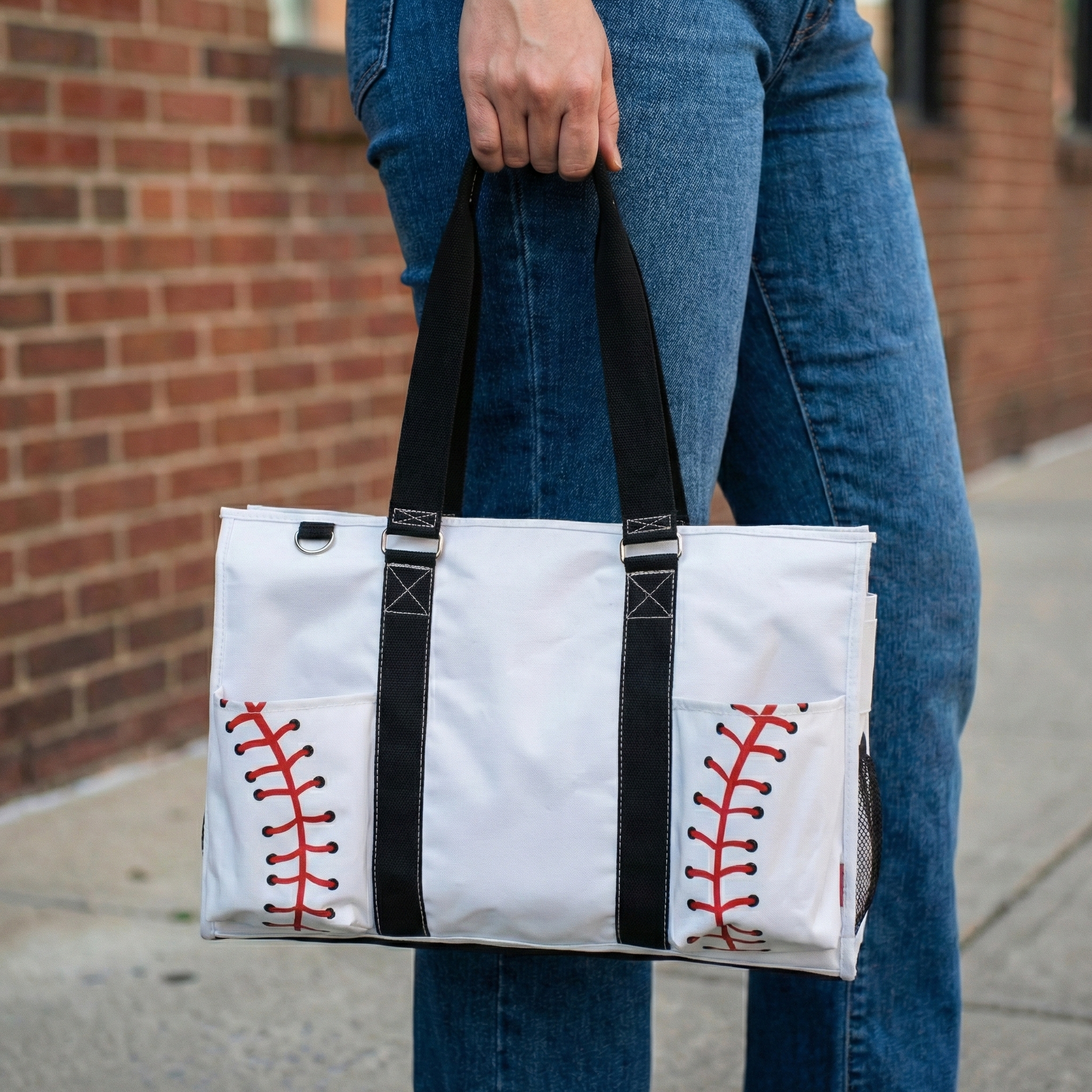 Person holding a white tote bag with black straps and red stitching, resembling baseball stitches, against a brick wall background.