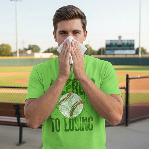 Man in a green t-shirt with a baseball graphic and text blowing his nose on a baseball field.
