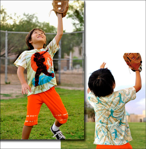 Child in a tie-dye shirt and orange shorts catching a baseball with a baseball glove.