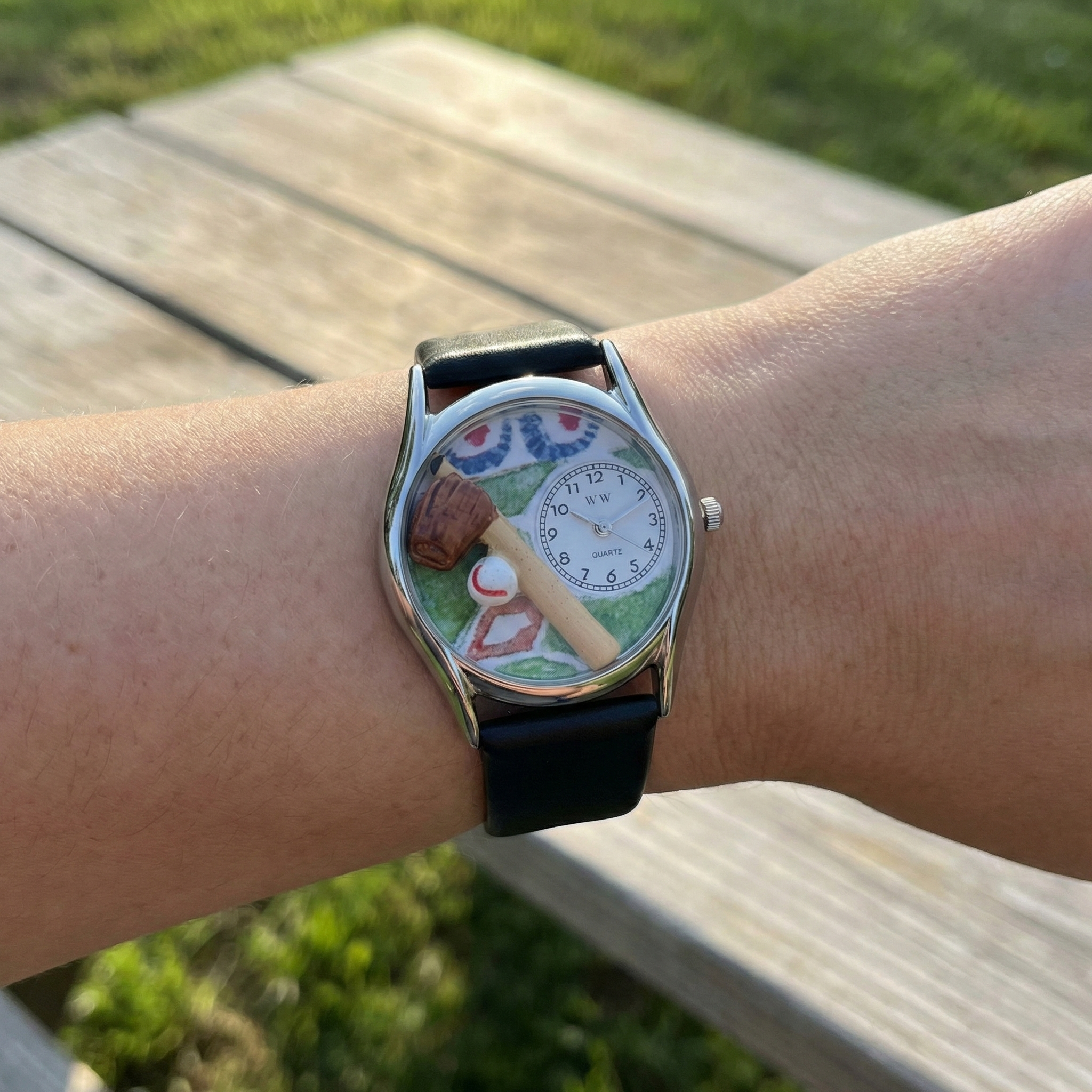 Wrist wearing a watch with a baseball-themed face, outdoors on a wooden bench.