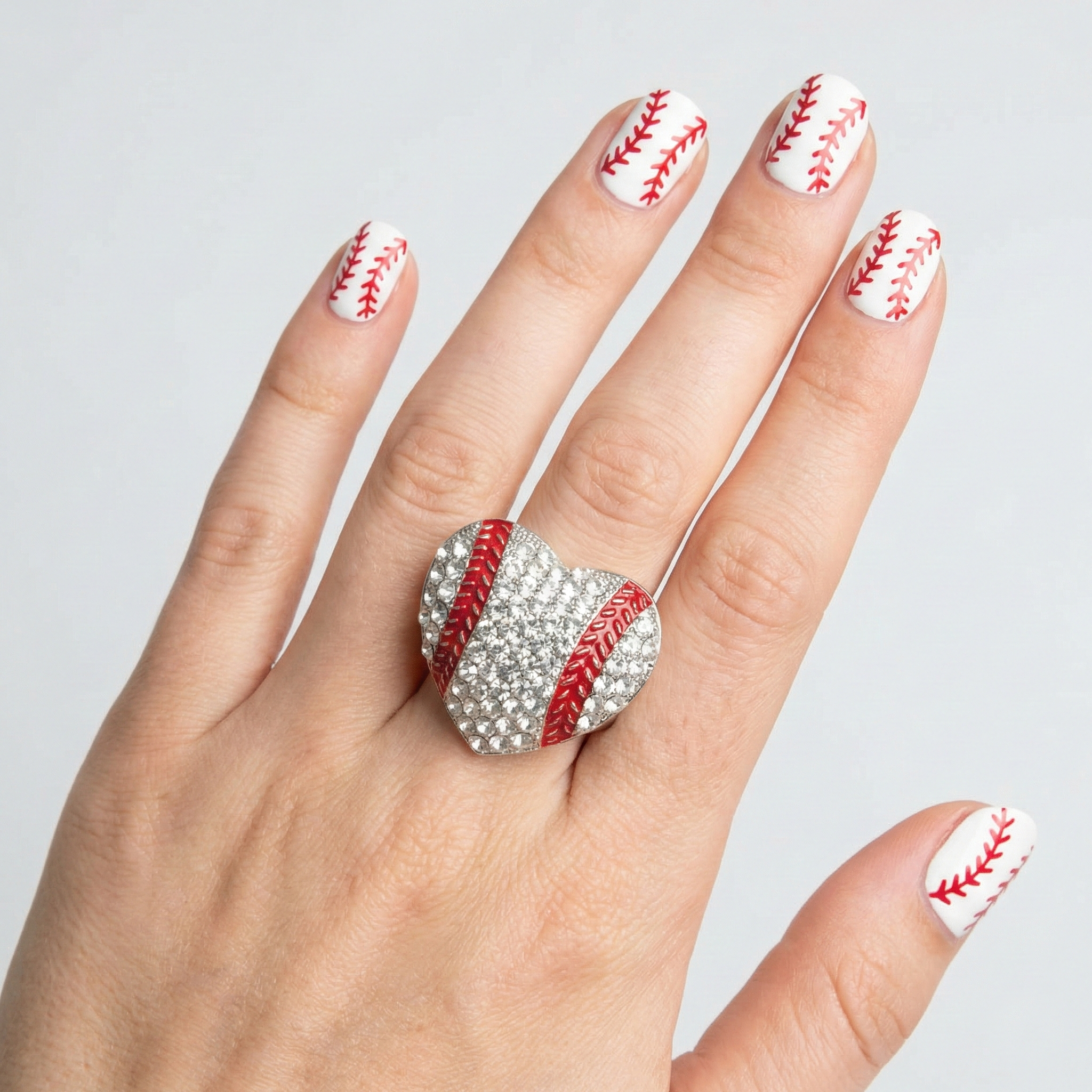 Hand wearing a baseball-themed ring with matching nail design on a light gray background