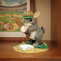 Stuffed animal rabbit playing baseball on a decorative base with a baseball and helmet, in front of a baseball-themed painting.