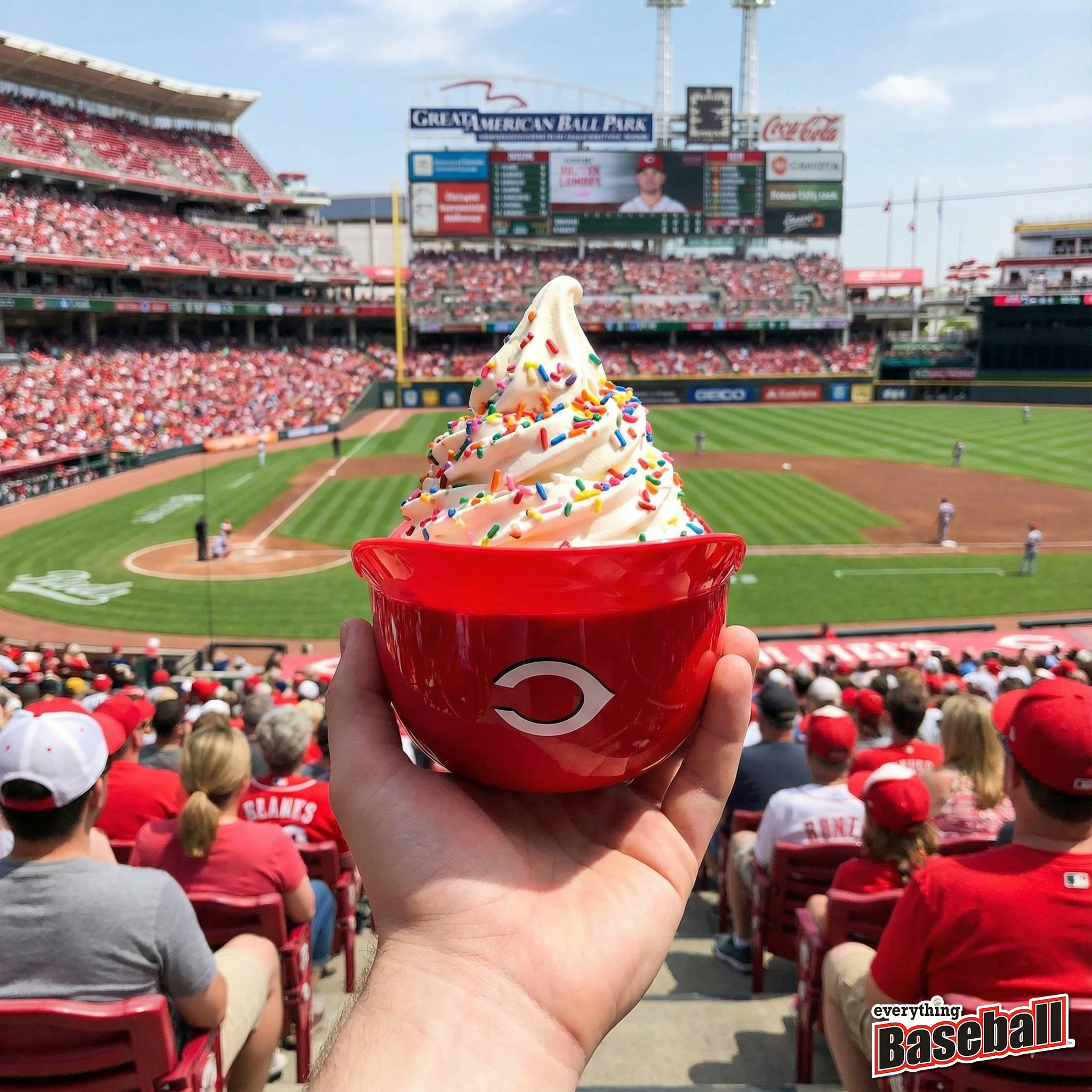 Hand holding a red Cincinnati Reds ice cream helmet bowl with colorful sprinkles at a baseball game.