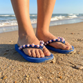 Blue baseball bead flip-flops with decorative straps worn on a sandy beach.