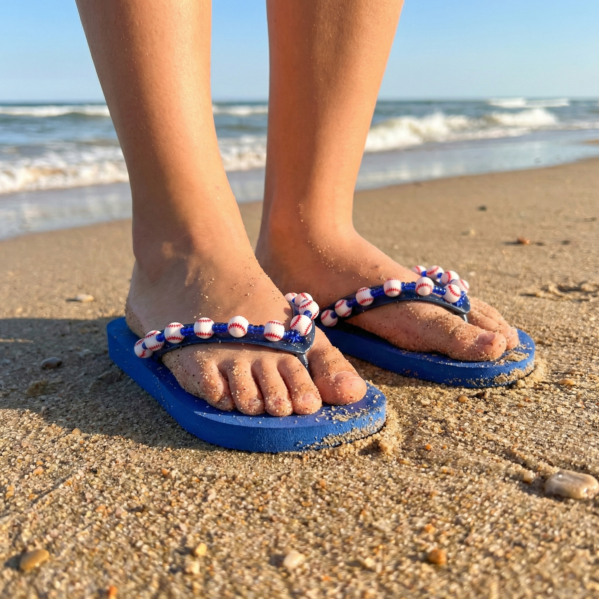 Blue baseball bead flip-flops with decorative straps worn on a sandy beach.