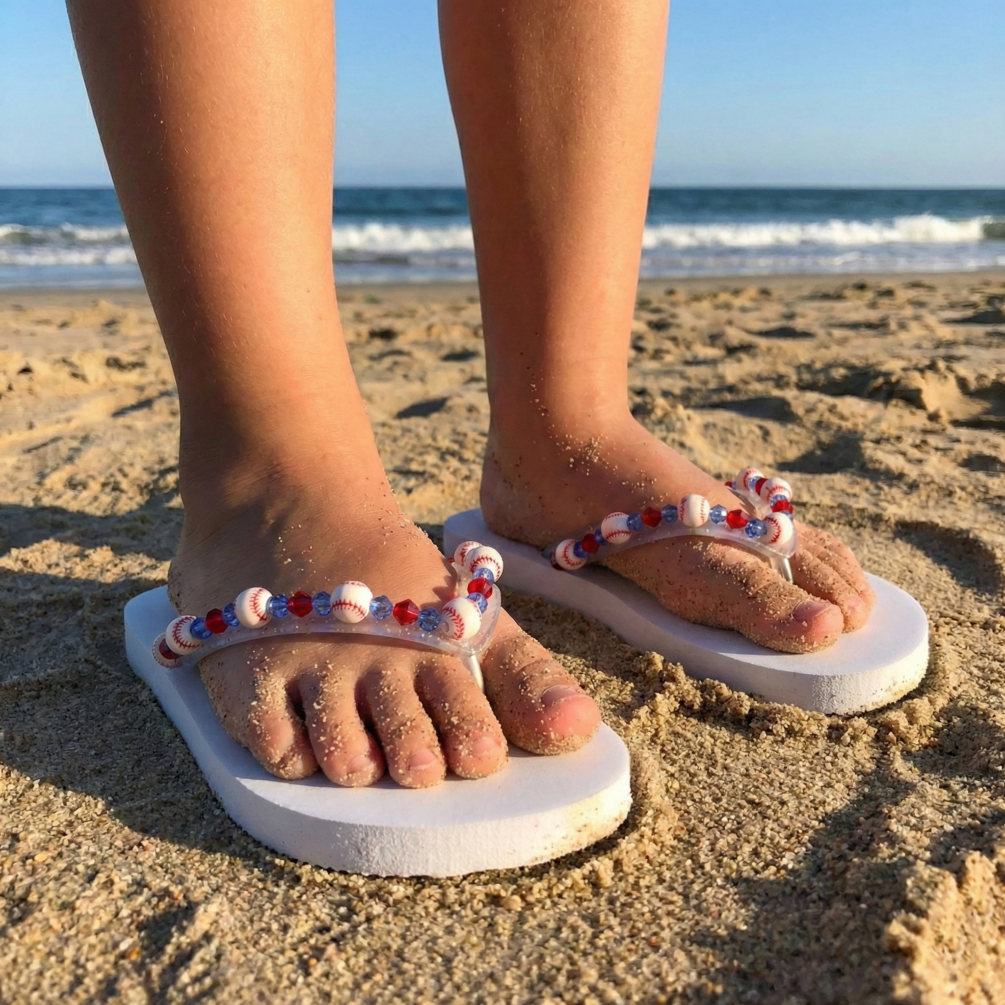 Feet wearing white sandals with decorative baseball bead straps on a sandy beach.