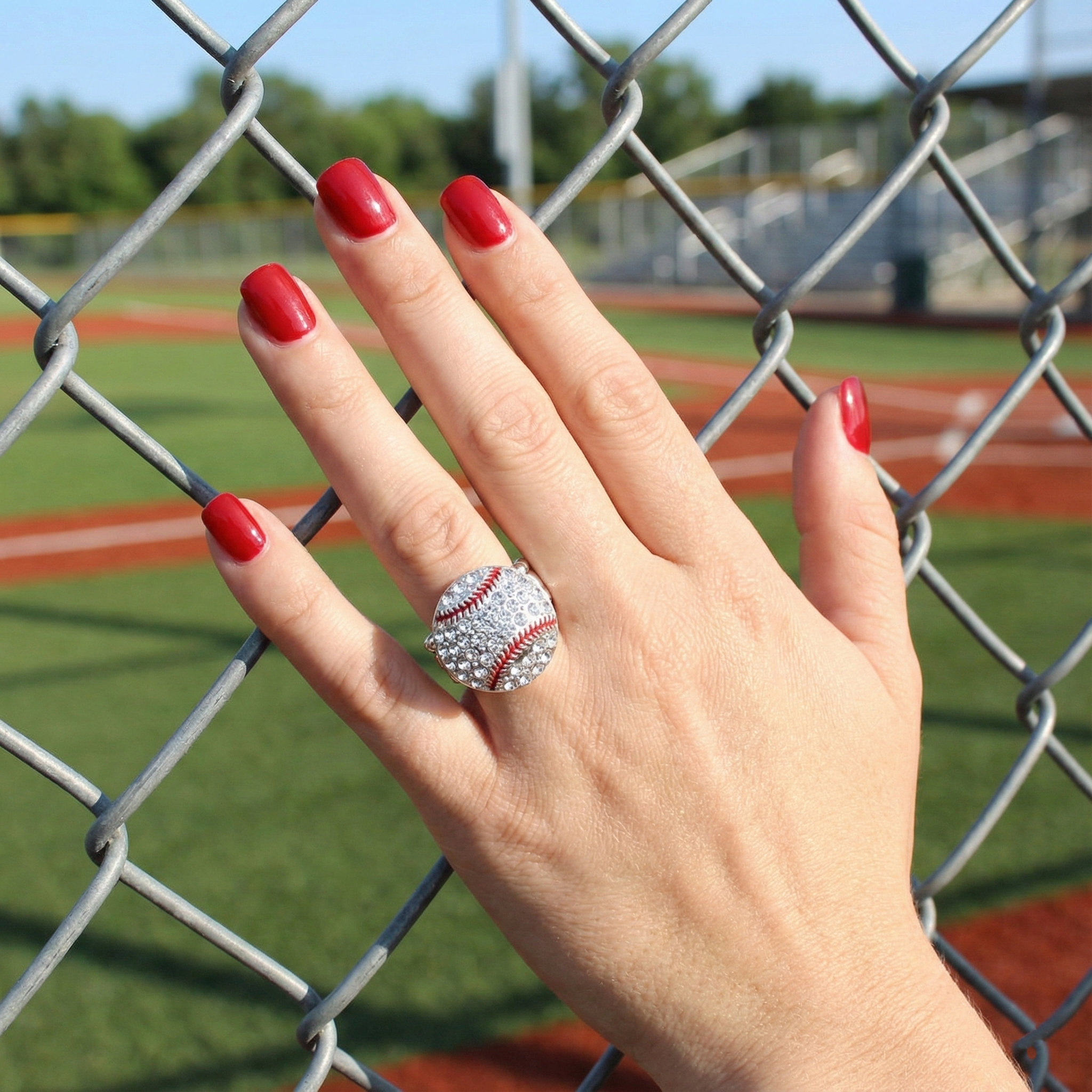 Hand wearing a baseball-themed ring with red nail polish against a chain-link fence background.